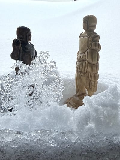 Two carved figures stand among snow and ice. The top figure, crafted from light brown wood, lies horizontally, partially buried in snow. Below, a darker figure, perhaps brown or black, is encased in a formation of clear, jagged ice crystals. The figures' shapes suggest human forms, possibly soldiers, with detailed carving showing clothing or armor. The background is obscured by more snow, creating a stark contrast with the figures and ice.