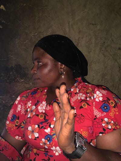 A woman wearing a black headscarf and a red floral top is shown in a close-up, sideways portrait. She has dark skin and is looking to the left. A silver cross earring is visible in her left ear. Her right hand is raised in front of her face, with her palm facing outward, and she is wearing a black wristwatch with a diamond-encrusted bezel. The background is a textured, dark wall. The image quality is moderately low, with visible grain and some motion blur on the hand. The scene appears to be indoors, possibly in a dimly lit room in Dull A, Nigeria.