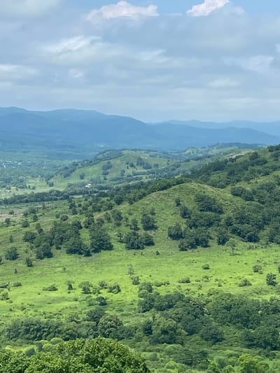The image displays a panoramic view of rolling green hills covered in lush vegetation and scattered trees. In the distance, a series of hazy blue mountains are visible, layered one behind the other. The sky is partly cloudy, with patches of white clouds against a pale blue expanse. The foreground is dominated by dense green foliage. A small, light-colored building with a dark roof is situated on a hillside in the mid-ground, surrounded by trees and fields. The scene suggests a natural, rural landscape, possibly in the vicinity of Kraskino, Russia, given the mention of the location. There are no people or animals visible in the image. The overall impression is one of serene natural beauty.