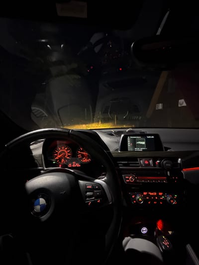The image shows the illuminated interior of a BMW car at night. The dashboard instruments are lit, showing the speedometer and other gauges. The climate control panel is visible, displaying a temperature of 20 degrees Celsius. The steering wheel, bearing the BMW logo, is prominently featured. The central display shows music information in Russian, with the artist "Я.Музыка". Through the windshield, a dimly lit urban environment can be seen, though it is difficult to make out specific details.
