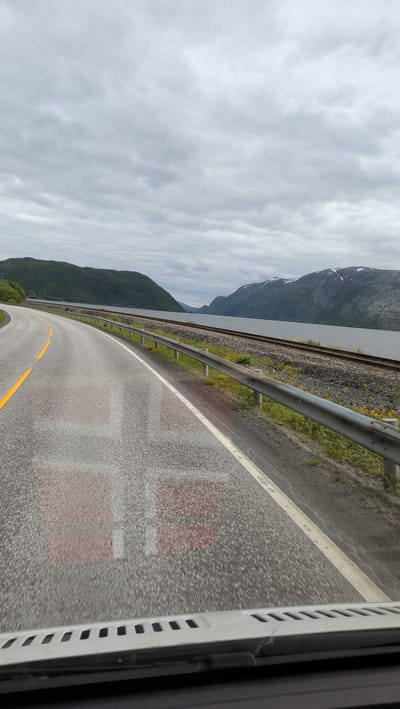 The image shows a road with a Danish flag painted on it. The road is curved and goes through a landscape with mountains and a body of water. The sky is cloudy. The image appears to have been taken from a vehicle, as the dashboard is visible at the bottom. The Danish flag on the road is faded and appears to be worn out. The location is identified as Mosjøen, Norway.
