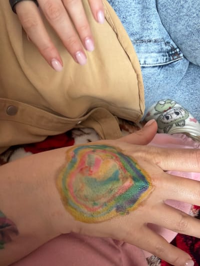 The image shows a close-up of a person's hand with a colorful, circular design on it. The design appears to be a temporary tattoo or body paint, featuring swirling patterns of yellow, green, blue, pink, and red. The hand is resting on a pink fabric. In the background, parts of blue denim clothing and a tan jacket are visible. The person's fingernails are painted a light pink color. There are no discernible elements in the image that would indicate a specific location within Valozhyn, Belarus.