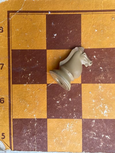 A close-up shot depicts a tan-colored chess knight piece resting on a vintage, checkered chessboard. The chessboard's squares alternate between tan and brown. The board's surface is worn and has signs of age, including discoloration and small white spots. The lower edge of the board shows numbers '3', '7', '6', and '5', each indicating the row of squares. The location in Zhaltusha, Bulgaria is unknown.