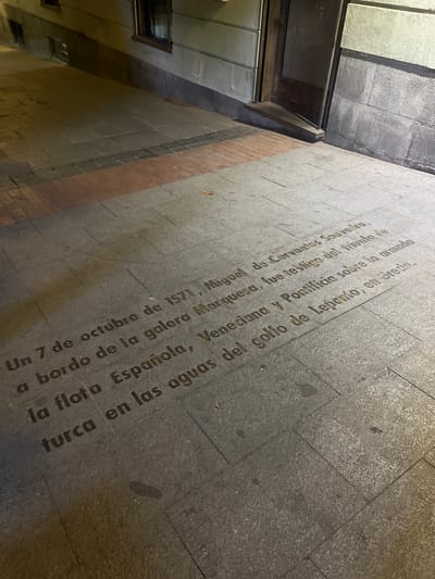 The image shows text inscribed on a stone sidewalk in Madrid, Spain. The text reads, "Un 7 de octubre de 1571, Miguel de Cervantes Saavedra, a bordo de la galera Marquesa, fue testigo del triunfo de la flota Española, Veneciana y Pontificia sobre la armada turca en las aguas del golfo de Lepanto, en Grecia." This translates to, "On October 7, 1571, Miguel de Cervantes Saavedra, aboard the galley Marquesa, witnessed the triumph of the Spanish, Venetian, and Pontifical fleet over the Turkish armada in the waters of the Gulf of Patras, in Greece." The sidewalk is made of large, rectangular stone tiles, with some darker tiles and red brick accents in the background. The scene is lit by artificial light, creating shadows on the sidewalk. The building facade behind the sidewalk is made of stone blocks and has a window and a doorway.