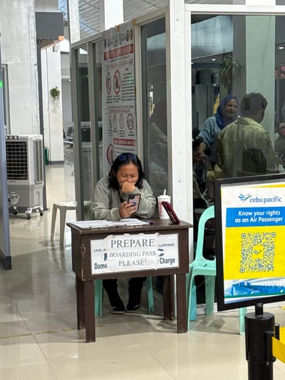 The image shows an indoor scene at an airport in the Philippines, likely in or near Del Carmen, based on the "ILIPPINES" text on the sign. A woman stands at a podium labeled "PREPARE BOARDING PASS PLEASE!" examining her phone. A cup of Coca-Cola sits on the podium near a pen. A sign advertising Cebu Pacific and air passenger rights is visible in the background. People are reflected in a window behind her. An air conditioning unit is located on the floor. Signs are posted with safety and prohibited items.