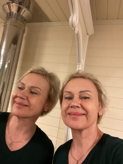 In the city of Korvet, Russia, a woman with short blonde hair and light blue eyes is reflected in what appears to be a polished metal flue pipe. She is wearing a dark green V-neck shirt and a gold necklace with a pendant. Her expression is a warm smile, with her head tilted slightly to her left. The background consists of light-colored horizontal wooden paneling and white decorative trim.