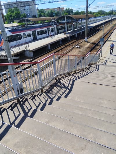 The image depicts a railway station in Monino, Russia, on a clear day. The foreground shows a set of concrete stairs with metal railings, casting long shadows from a railing onto the steps. The stairs lead down towards a railway platform. In the middle ground, a train with white, red, and blue livery is stopped at the platform. Several passengers are visible on the platform, some standing near the train doors. The railway tracks run alongside the platform. In the background, trees and apartment buildings are visible under a bright blue sky with scattered clouds. A man in a blue shirt and dark pants is walking away from the camera on the platform in the middle distance. Another man in a light-colored shirt and dark pants is walking on the platform further down the tracks.