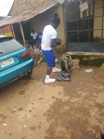 In Ohit, Nigeria, a young man wearing a white t-shirt, blue shorts, white socks, and white sneakers stands on a dirt and gravel surface next to a teal car. He is holding a black garment in his hands and looking down. To his right, a beige backpack rests on the ground next to a folded dark-patterned cloth with a phone on top. In the background, two other young men in white t-shirts and blue shorts sit on a bench under a corrugated metal roof attached to a building with a brick and stucco exterior. A black metal grate covers a window or doorway behind them. The car, a Honda Civic, has a license plate that reads "BC137-KMC". The environment appears to be a roadside or outdoor market area, with debris and puddles visible on the ground.