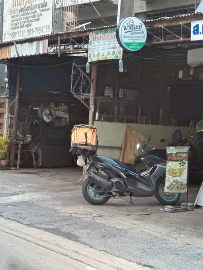 The image shows a street scene in the city of Nonthaburi City Municipality, Thailand. In the foreground, a black motorcycle with a teal-colored rim is parked on a cracked asphalt surface. Attached to the rear of the motorcycle is a bright orange delivery box. To the right of the motorcycle, a white stand displays a food menu with pictures of dishes and Thai text.
Behind the motorcycle, a covered outdoor area with a corrugated metal roof is visible. A large orange delivery bag is suspended from a rack in this area. Further back, there are structures with wooden supports and corrugated metal siding. Potted plants are present near the entrance to one of the structures. Various signs are displayed, including a circular green and white sign with Thai text and a smaller rectangular sign with a food menu. The overall environment suggests a commercial area, possibly a restaurant or food stall.