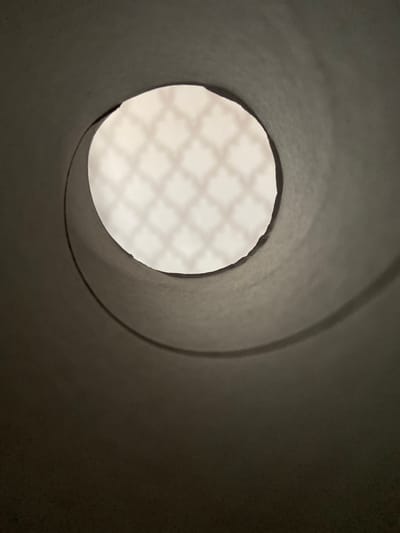 The image shows a close-up, low-angle view looking up through the hollow center of a cardboard tube, possibly a toilet paper roll or paper towel roll. The inside of the tube is a dark, matte grey, with a spiral shadow and texture visible on the inner surface, suggesting the roll has been partially used or is slightly unraveled. The opening of the tube reveals a bright, diffused light source beyond. The light filters through a translucent material with a faint, repeating geometric pattern, resembling a diamond or lattice shape. The overall impression is one of looking through a tunnel or aperture towards a softly illuminated background. There are no people or discernible objects within the immediate scene. The image was taken in Beloretsk, Russia.