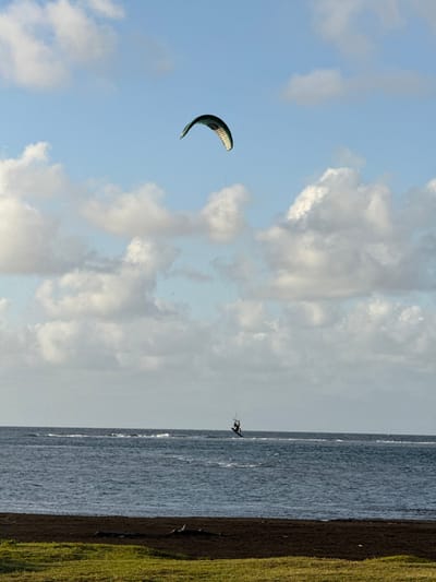 The image depicts a kitesurfer active on the water in Māhina, France. In the upper sky, a dark green and light-striped arched kite is visible, fully inflated and aloft, connected by lines to a single individual on the water below. The kitesurfer, wearing dark attire, is positioned on a board, creating a wake across the dark blue-gray water surface. The horizon separates the water from a partly cloudy blue sky, featuring scattered white and gray cumulus formations. The immediate foreground comprises a strip of dark, possibly volcanic, soil or sand, containing several unidentifiable dark objects, bordered by a patch of green grass.