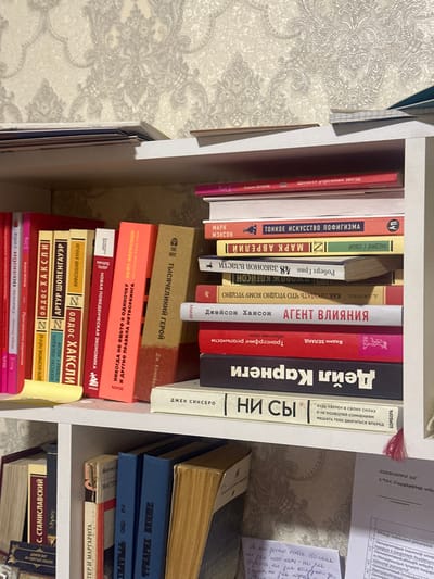This image depicts a section of a white wooden bookshelf inside a room in the city of Tairov, Armenia. The background features patterned wallpaper with a light beige or cream base and a slightly darker, ornate damask-like design. The top of the visible shelf unit holds a stack of papers or magazines on the left.
The upper shelf contains numerous books. On the left side, a row of books stands vertically, predominantly with red, yellow, orange, and light brown spines. Notable visible titles, primarily in Cyrillic script, include "Олдос Хаксли," "Артур Шопенгауэр," "Никогда не ешьте в одиночку," and "Тысячелетний герой." A yellow sticky note is partially affixed to one of the red book spines on the far left. On the right side of this shelf, a vertical stack of horizontally placed books is visible. From top to bottom, prominent titles include "Шелест утренних звезд," "Марк Мэнсон / Тонкое искусство пофигизма," "Марк Аврелий," "Роберт Грин / 48 Законов Власти," "Джейсон Хэнсон / Агент влияния," a red book with "Трансерфинг реальности," "Дейл Карнеги," and "Джен Синсеро / Ни сы."
The lower shelf, partially visible at the bottom of the image, also holds books standing vertically, including "К. Станиславский" and "Мастер и Маргарита," alongside several blue-bound volumes. To the right of the lower shelf's books, loose papers are present; one appears to be a document with Armenian script at the top, and another contains handwritten text in Cyrillic. A red tassel hangs down from the upper shelf, resting near these papers. No individuals are present in the scene, and no specific actions or interactions are depicted beyond the arrangement of objects.