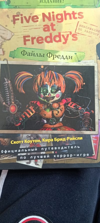 The image shows the cover of a Russian-language book titled "Five Nights at Freddy's: Freddy Files" by Scott Cawthon and Kira Bridd-Raisly. The cover features a large image of a robotic character from the Five Nights at Freddy's game series. The character has orange hair, a light green face with red circles and dark green eyes, and is wearing a red and orange metallic outfit with exposed wires and gears. The character is holding a large, red, metallic object in its right hand. The background of the image is black. The book's title, authors' names, and tagline "Official Guide to the Best Horror Game" are written in Cyrillic text on a black and yellow background. The image is cropped at the bottom, showing a small portion of a black garment. This could be a book cover, a book in a bookstore, or a book on a table or shelf. The image was likely captured in Ganja, Azerbaijan.