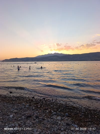 The image depicts a serene sunset scene over a body of water, presumably a lake or sea, with a mountainous coastline in the distance. The sky is a gradient of soft blues, oranges, and pinks, with sunbeams radiating from behind the mountains. A few scattered clouds are tinged with the warm hues of the sunset.
Several people are visible in the water, engaged in swimming and wading. Some are closer to the shore, while others are further out. There appears to be a jet ski in the middle distance, indicating recreational activity. Buoys or markers are also scattered across the water's surface.
The foreground shows a pebbled beach with some sparse vegetation. The water is calm, with gentle waves lapping at the shore. The overall atmosphere is peaceful and idyllic, characteristic of a summer evening. The image is watermarked with "REDMI NOTE 13 PRO" and a timestamp "27.06.2026 19:42".
While the specific location of Yusuphana, Uzbekistan is mentioned in the prompt, there are no visual elements in the image that definitively confirm or deny this location. The landscape with mountains and a large body of water is consistent with various geographical settings.