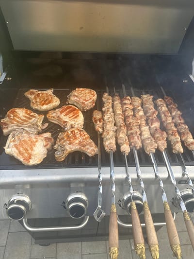 Six pork chops and eight skewers of meat are grilling on a stainless steel barbecue. The pork chops are arranged on the left side of the grill, some showing char marks from the heat. The skewers of meat, likely kebabs, are on the right side of the grill, with the meat appearing browned and slightly rendered. Smoke rises from the grill, indicating the cooking process is active. The barbecue has two control knobs with labels for "HIGH" and "OFF," and a silver handle across the front. The background shows the inside of a grill lid, which is partially open. The scene takes place outdoors, with a hint of paved ground visible at the bottom of the frame. The location is identified as Targai, Russia.