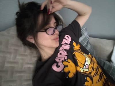 An indoor scene in Rasskazovo, Russia, depicts a young adult female from a slightly elevated perspective. The subject is lying on her back, angled with her head towards the upper right of the frame. She possesses dark, medium-length hair, styled with bangs, and wears dark-rimmed eyeglasses. A small stud earring is visible in her left ear. Her lips are pursed, and her gaze is directed away from the camera. Her right hand, adorned with dark red nail polish, is positioned above her head, gently touching her hair. She is wearing a black short-sleeved t-shirt with a pink "GARFIELD" text logo and a large orange Garfield character printed on the front. She is resting on what appears to be a light brown, subtly textured upholstered surface or blanket on the left, and a gray plaid patterned fabric, likely a pillow or blanket, on the right. A plain, light gray wall or headboard serves as the background behind her head. The image exhibits a soft focus, with slight blur visible on the lower left portion of the frame.
