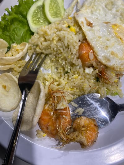 A plate of nasi goreng (fried rice) is served with a fried egg, two cooked shrimp, lettuce, cucumber slices, and prawn crackers. A fork and spoon are placed on the plate, indicating that the meal is being eaten. The image was taken up close, focusing on the food. The setting is an indoor environment with a white plate and table. The food appears to be prepared and ready for consumption.