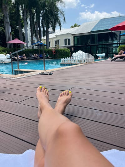 In Bang Sao Thong Subdistrict, Thailand, a person's legs, with bright yellow toenails, are prominently featured in the foreground, resting on a wooden deck by a swimming pool. In the background, the pool area is visible, with lounge chairs, umbrellas, and several people relaxing. Some individuals are in the pool, while others are lying on the chairs. The scene is set against a backdrop of tropical vegetation and modern buildings, under a partly cloudy sky.