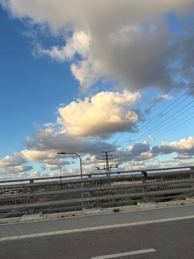 The image depicts a static scene from an overpass or bridge in Hofit, Israel, under a partially cloudy sky. The sky features distinct patches of bright blue and numerous cumulus clouds, varying in brightness with areas illuminated by sunlight and others in shadow.
In the immediate foreground, a grey metal guardrail system, composed of horizontal bars and vertical posts, is mounted on a concrete barrier. To the right of the guardrail, a paved pedestrian walkway is visible, constructed from grey and white interlocking pavers. Adjacent to the walkway on the left, an asphalt road surface is present.
From the left side of the bridge, a tall street light with a curved arm and a dark, horizontal fixture extends upwards. In the mid-ground, a large metal electricity pylon, characterized by its lattice structure, is situated on the right side, supporting multiple overhead power lines that traverse diagonally across the upper right portion of the sky. A dark, unidentified object is suspended from the guardrail on the right side of the bridge.
Below the bridge, a multi-lane roadway is visible, featuring white lane markings and bordered by sparse vegetation. No individuals, vehicles, or dynamic actions are present in the scene.