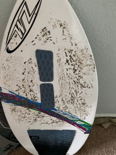 A white wakeboard is leaning against a textured, light blue wall. The wakeboard has a graphic logo on the upper left that reads "WL" in a stylized font within an oval. The board is dirty with a significant amount of dark, sandy debris scattered across its surface. There are three traction pads on the wakeboard: two rectangular pads stacked vertically in the center and one larger pad on the bottom. The top two pads are dark gray with a diamond pattern. The bottom traction pad is also dark gray with a diamond pattern and has a black logo that appears to say "DK". A colorful stripe design, featuring lines of purple, blue, and green, is painted on the wakeboard near the bottom. The wakeboard is positioned indoors, on a carpeted floor, next to a beige fabric strap. The background suggests an indoor environment, possibly a home. No individuals are visible in the image. The wakeboard appears to be at an address in Elizabeth City, United States.