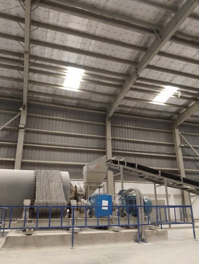 The image shows a large industrial facility in Pangasia, Bangladesh (latitude: 22.694, longitude: 90.601). The scene is a large, open industrial space with a metal roof and corrugated metal walls.  In the foreground, there is a large, industrial-sized machine, with a large, cylindrical drum, a conveyor belt, and an electric motor. The machine is secured behind a blue, metal safety railing. The metal roof is supported by large, metal beams. The floor is concrete and dusty. There are no people in the image. The image was taken indoors in a brightly lit, industrial setting.