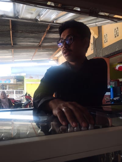 The image depicts a medium shot of a young man wearing glasses and a black shirt, seated indoors. He is looking to his right, with his left hand resting on a glass surface that reflects a row of small, bottle-like objects. A ring is visible on his left ring finger. In the background, a woman wearing a headscarf is visible to the left, and a red motorcycle is parked behind her. The environment appears to be a shop or business, with a corrugated metal roof and some signage visible in the distance. The overall scene suggests a casual, possibly commercial setting in Pompanua, Indonesia.