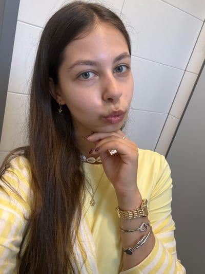A young woman poses for a selfie in what appears to be an indoor location with white, rectangular tiles visible on the wall behind her. She is wearing a yellow top with white stripes. Her dark brown hair is long and covers part of her shoulder. On her wrist, she is wearing a gold watch and two silver bracelets. She has fair skin and blue eyes. The location and city in Germany, Hanau, are unknown.