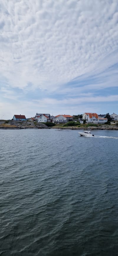 The image depicts a coastal scene in Öckerö, Sweden, under a partly cloudy sky. In the foreground, choppy, dark blue water fills the lower half of the frame. A white motorboat is visible in the mid-ground, moving from left to right and leaving a wake. In the background, a rocky shoreline is lined with several houses, most of which are white with red roofs, characteristic of Scandinavian architecture. Some houses are two stories, and they appear to be situated on a slight elevation. The land is dotted with green vegetation. The sky above is a mixture of blue sky and scattered white, textured clouds.