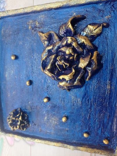 The image shows a close-up of a textured blue surface with gold accents. A prominent, dark blue rose with gold highlights is in the center-right of the frame. The rose has many layered petals and appears to be a sculpted or raised element on the surface. Scattered across the blue background are several small, raised gold spheres, resembling beads or decorative dots. To the lower-left of the main rose, there is a smaller, similarly textured dark blue and gold floral element. The edges of the object appear to have a rough gold finish. The overall aesthetic suggests a decorative art piece or craft item. The location "Дукорскі сельскі Савет, Belarus" is irrelevant to the visual content of the image.