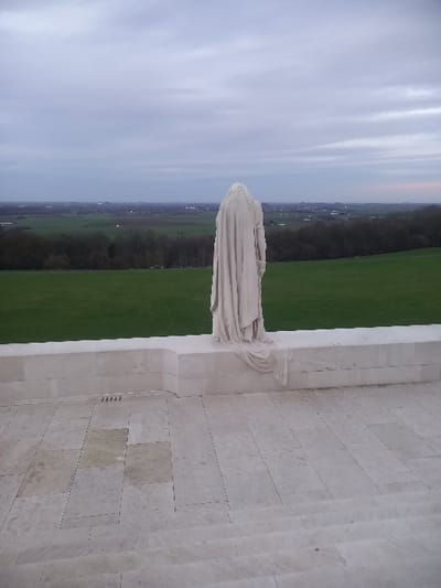 A statue of a draped figure stands atop a light-colored stone structure. Beyond the monument stretches a landscape of vibrant green fields bordered by a dark strip of trees, possibly a forest or grove. The sky above is overcast with muted light. The ground appears to be constructed of horizontal stone blocks.