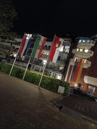 The image captures a night scene in Emmerich am Rhein, Germany. Four flags are prominently displayed: the Dutch flag (red, white, and blue), the flag of North Rhine-Westphalia (green, white, and red with the coat of arms), the flag of Emmerich am Rhein (red and white with a bucket), and the German flag (black, red, and gold). The flags are mounted on poles and stand in front of a green hedge. Behind the flags are buildings, with windows visible. The foreground shows a paved area. The overall lighting is dim, suggesting it is late evening or night. No people are visible.