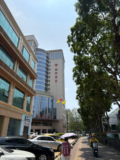 The image captures an upward perspective of a vibrant street scene in 前埔村, China, under a clear, bright sky during the daytime. Lush green trees with dense foliage form a natural canopy across the top of the frame, shading the sidewalk below.
Prominent in the background is a tall, modern building featuring a light-colored facade adorned with bold red Chinese characters, "厦门国际银行" (Xiamen International Bank), and a distinctive red circular logo. Below this, the structure transitions into a section characterized by blue-tinted glass windows with a sweeping, curved design. In the foreground, another building displays large, reflective blue-tinted windows set within brown, rectangular frames.
Along the paved sidewalk on the right, several cars are parked, including white, black, and silver models. A white umbrella and a purple suitcase are visible near the parked vehicles. A person in a yellow uniform and helmet, likely a delivery driver, is seen riding a motor scooter on the sidewalk, with other scooters further down the street. Decorative yellow and red flags hang from the Xiamen International Bank building. Visible text on storefronts includes the English word "DunDun" and Chinese characters such as "家佳乐" (JiaJiaLe) and "福记" (Fuji), indicating various businesses. The scene portrays a typical urban environment with active daily life.