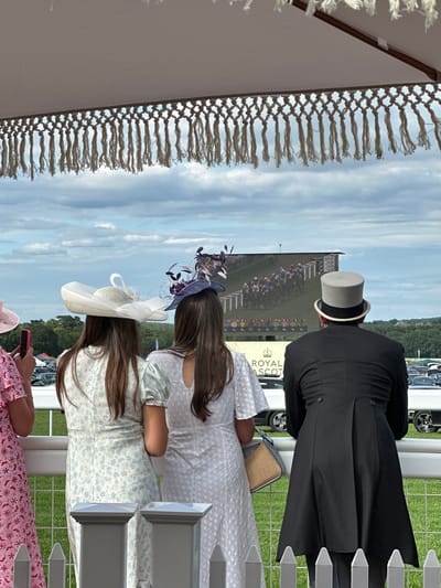 A group of people are watching a horse race on a large screen at Royal Ascot in Ascot, United Kingdom. The screen shows a field of horses with jockeys in colorful silks, racing on a track. The people are dressed in formal attire, including hats and suits, befitting the event. One person in the foreground wears a black tailcoat and a grey top hat. Two women to the left are wearing elaborate hats and dresses. The scene is set outdoors, with a cloudy sky and green fields visible in the background. The event appears to be a social gathering with a focus on the horse racing spectacle.