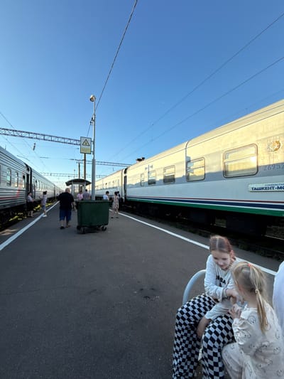 The image depicts a train station platform, possibly in Uzunovo, Russia, under a clear blue sky. Two trains are present, one on the left and another on the right, running parallel to the platform. A group of people, including adults and children, are visible on the platform. Some individuals are standing near the trains, while others are seated. Two young girls are sitting on a bench in the foreground, facing each other and interacting. One of the girls is wearing checkered pants and a white long-sleeved shirt, while the other is in pajamas with a pattern of cartoon characters. A green trash bin is located on the platform between the two trains. Overhead, power lines and a lamppost are visible, along with a warning sign. The platform itself is paved and has a white line marking its edge. The lighting suggests it is either early morning or late afternoon.