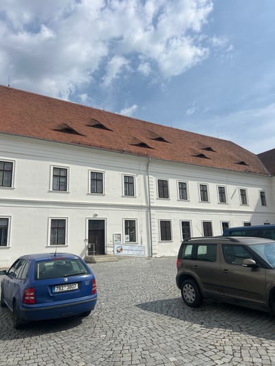 This media file captures a street scene in Rosice, Czechia, featuring a white building with a red-tiled roof and several cars parked on a cobblestone street. The building appears to be a historical structure, possibly a school or a government building, given its grand architecture and the multiple windows arranged in neat rows. Some of the windows have dark shutters. The roof is steeply pitched and covered with terracotta tiles. A few dark, diamond-shaped dormer windows protrude from the roof.
Two cars are visible: a blue hatchback with the license plate "7B2 3807" and a silver-brown station wagon. The blue car is parked closer to the building, while the station wagon is further down the street. The cobblestone street suggests an older, traditional European town.
The sky is blue with scattered white clouds, indicating a partly cloudy day. The sun is casting shadows, suggesting it is daytime, likely late morning or early afternoon. There are no people clearly visible in the image.
A sign is partially visible on the building, with some text in Czech. It appears to be a directory or information board. The overall impression is one of a calm, sunny day in a historic European town.