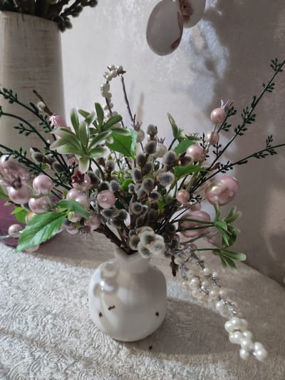 A close-up shot captures an Easter-themed floral arrangement in a white ceramic vase. The arrangement features delicate pink faux berries, fluffy catkins, and sprigs of greenery. A strand of faux pearls drapes from the bouquet onto a textured white tablecloth. In the background, a portion of a light-colored vase and a decorative egg are visible against a textured wall. The scene is likely set indoors, possibly in a home, given the decorative elements and domestic setting. The image does not contain any information that would allow for the determination of its location in Akhalkalaki, Georgia.