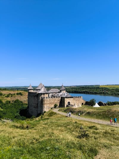 The image depicts the Khotyn Fortress, situated on the right bank of the Dniester River in Khotyn, Chernivtsi Oblast, Ukraine. The scene is observed on a clear, sunny day with a deep blue sky dominating the upper half of the frame.

The Khotyn Fortress, constructed from stone, features multiple turrets with conical wooden roofs and crenellated battlements. The fortress complex itself is positioned on a raised landform adjacent to the river, with its lower section partially obscured by green vegetation. A winding dirt path, visible in the foreground, leads towards the fortress.

Several individuals are present on the path. Two adults and one child walk closer to the fortress, while another group of three individuals (two adults and one child) walk further down the path towards the viewer. Their attire is casual. Two small, light-colored, conical tents are pitched on the grassy area near the fortress walls.

The Dniester River flows behind the fortress, reflecting the bright sky. The opposite bank of the river is characterized by dense green foliage and agricultural fields under the clear sky. The immediate foreground consists of a gently sloping hill covered in dry and green grasses, with some sparse bushes.
