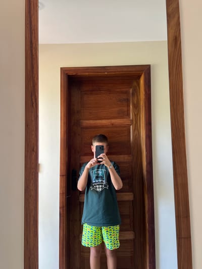 The image shows a young male taking a self-portrait in a full-length mirror within an indoor setting in Michamvi Kae, Tanzania. The male, with short hair, wears a dark green t-shirt featuring a light-colored crest logo and bright green shorts adorned with a repeating pattern. A silver watch is visible on his left wrist. His face is entirely obscured by a black smartphone, held horizontally with both hands at chest level, directed towards the mirror to capture the reflection. The mirror features a thick, dark reddish-brown wooden frame with visible grain. Behind the male's reflection, a large, dark reddish-brown paneled wooden door with matching trim is prominent, flanked by a light cream-colored wall section. A white ceiling is visible above the door. The scene is well-lit, characteristic of a room within a building, likely a hotel or residence.