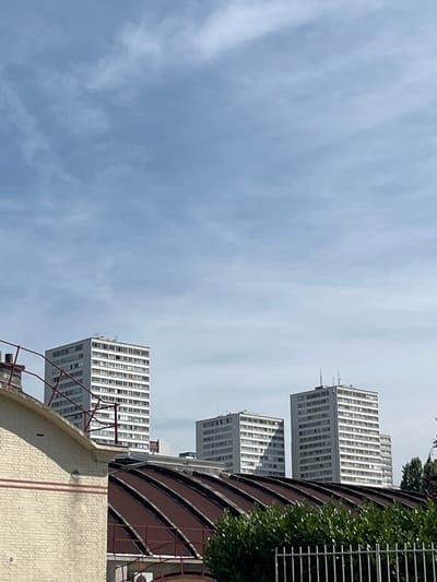 This is a medium shot of three tall white apartment buildings under a bright, partly cloudy blue sky. The buildings are arranged in a line, with the tallest and most central one slightly further back than the other two. They are modernist in design, with many windows on each floor. In the foreground, a lower, cream-colored brick building with a red metal railing is partially visible on the left. A dark, curved metal roof extends across the lower part of the frame, obscuring the base of the apartment buildings. Dense green foliage is visible in the lower right corner, and a white metal fence is in the extreme foreground. The scene appears to be an urban residential area in Le Kremlin-Bicêtre, France, likely on a clear day. There are no people visible in the image.