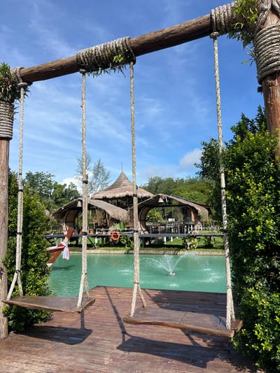 This image depicts a serene outdoor scene, likely a resort or restaurant, in Tambon Pa Kae Bo Hin, Thailand. In the foreground, two rustic wooden swings hang from a sturdy wooden beam supported by thick, rope-wrapped posts. The swings are suspended over a wooden deck. Beyond the deck, a vibrant turquoise body of water is visible, with a fountain spraying water in the center. In the background, several thatched-roof structures with open sides, typical of tropical architecture, are built over the water or on stilts. Lush green foliage and trees surround the area, and a clear blue sky with scattered clouds completes the picturesque setting. A small orange life preserver is hanging on a railing in front of one of the structures.