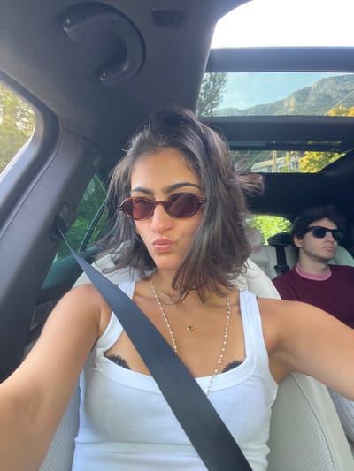 A woman wearing sunglasses and a white tank top, with a seatbelt across her chest, is taking a selfie inside a car. She is wearing a pearl necklace, and a tattoo is partially visible on her chest. Behind her and to the right, a man wearing sunglasses and a burgundy shirt is reclined in the passenger seat. The car's interior is predominantly black, with light-colored leather seats. Outside the car window, a mountain is visible, and greenery can be seen. The location is Èze, France.