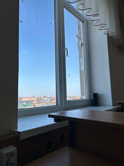 The image displays an interior perspective looking out through a multi-pane white PVC window, presumably located in Dmitrievskaya, Russia. The window glass is characterized by numerous small specks and multiple adhesive star-shaped decorations. The exterior view through the window reveals a clear blue sky in the upper portion, transitioning to a distant suburban landscape featuring a collection of houses with varying roof colors, including green and red, and sparsely treed or bare-branched vegetation indicative of a cool season. The window frame on the right side exhibits significant damage, with sections of plaster or concrete peeling away, exposing underlying brick or structural material.

Within the interior, a white window sill runs across the base of the window. On the right side of the sill rests a dark, rectangular object, possibly a block. In the foreground, a light-brown wooden desk or table surface is partially visible, supported by a black metal frame and secured with fasteners topped with white caps. Below the desk surface, a darker wooden panel includes two circular cutouts. To the far left, a white double electrical outlet is mounted on the wall below the window sill. On the right, a translucent curtain with a distinctive wavy brown trim element hangs vertically. No individuals are present in the scene, and no explicit actions or interactions are depicted. The overall environment suggests an institutional or residential room overlooking a populated area.