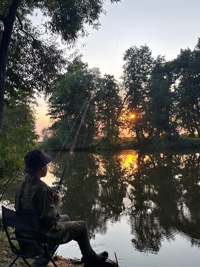 The image depicts a serene fishing scene at dusk in Boriskino, Russia. A young person, dressed in camouflage attire and a bucket hat, is seated on a foldable chair by the edge of a calm body of water. They are holding a fishing rod, actively engaged in fishing. The setting sun casts a warm, golden glow on the scene, illuminating the trees on the opposite bank and their reflections in the water. The sky is a soft gradient of orange and pale blue. The overall atmosphere is tranquil and peaceful.