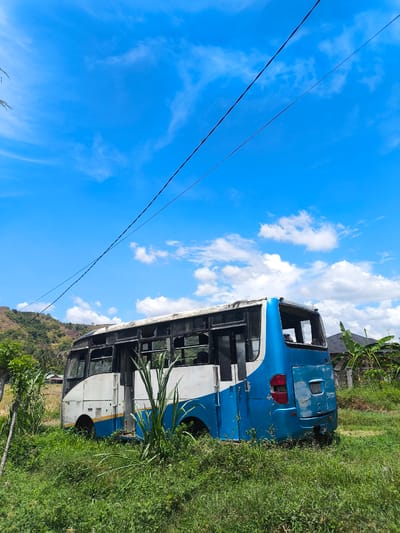 An abandoned blue and white bus sits in a grassy field under a bright blue sky with scattered white clouds. The bus is weathered and appears to be missing windows and parts of its roof. Vegetation grows around and within the bus. In the background, a green hillside with trees and a small building can be seen. A power line stretches across the top of the frame. The location is Meunasahjok, Indonesia.