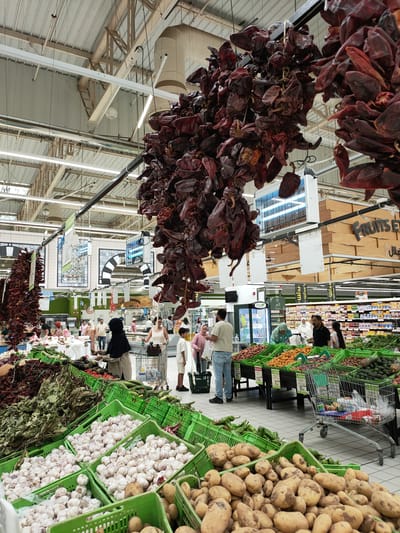 The image depicts the interior of a supermarket in Cebalat, Tunisia. In the foreground, several crates overflow with fresh produce, including large quantities of garlic bulbs and potatoes. Beyond these, a variety of fruits and vegetables are displayed in green crates, such as apricots, peaches, kiwis, and cucumbers. Hanging above the produce sections are clusters of dried red peppers, adding a rustic element to the market.

In the middle ground, shoppers navigate the aisles. A woman wearing a dark headscarf is seen interacting with produce, while other individuals, including a man in a black polo shirt and a young boy, are also present. Shopping carts are visible, indicating ongoing purchases.

The background reveals the typical supermarket environment with shelves stocked with packaged goods, overhead lighting, and various signs, some featuring Arabic script. The overall scene suggests a bustling marketplace where fresh produce is a central offering.
