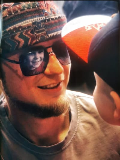 The media file is a close-up photograph of two people. The primary subject is a man wearing a patterned beanie and sunglasses. His reflection in the sunglasses shows a young child smiling. The man has a light beard and is wearing a grey t-shirt. To the right of the man, a portion of another person wearing a red and white hat is visible. The lighting suggests it might be daytime, possibly outdoors given the reflection of a child in sunglasses. The overall emotion conveyed is one of warmth and connection, likely a family moment. No text is visible in the image.