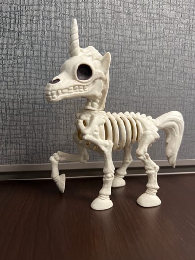 The image shows a white skeleton toy unicorn standing on a dark wooden surface, with a grey textured wall in the background. The unicorn is depicted in a side profile, facing left. Its body is made of white plastic, mimicking the appearance of bones, with visible ribs, spine, and leg structure. The unicorn has a single horn on its forehead, large black eye sockets, and a skeletal jaw. Its tail is flowing, and one of its front legs is slightly raised. The lighting suggests an indoor environment, likely a desk or shelf, given the dark wood surface and the wall texture which resembles fabric or a bulletin board. There are no people or discernible city elements present in the image. The location is not specified within the image itself, and therefore, its presence in Columbia, United States of America cannot be confirmed.