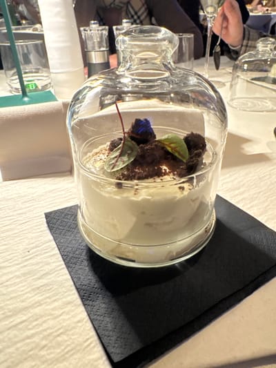 The image displays a creamy, light-colored dessert served in a clear glass container covered by a matching glass cloche. The dessert is topped with dark brown crumbles, a dusting of cocoa powder, two small green leaves, and a small dark blue/purple item. The container rests on a black, textured paper napkin, which is situated on a white, textured tablecloth. In the background, a dining setting is visible within a restaurant in Bolzano, Italy. To the left, a folded white napkin, a green utensil, and a clear drinking glass are present. A salt and pepper shaker set stands further back. On the right, a second identical glass cloche is partially visible. Blurred figures of diners are in the background; one individual's hand, wearing a plaid sleeve, is seen holding a spoon or fork.