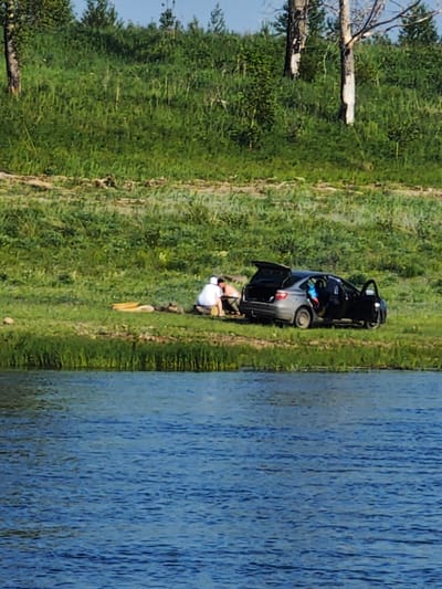 The image depicts a scene beside a body of water, likely a river, with a grassy embankment leading up to a wooded area. In the foreground, the water is a deep blue with visible ripples. On the grassy bank, a grey sedan is parked with its rear hatch and driver's side door open. Two individuals are present near the car. One, wearing a white top and dark pants, is seated on the ground, facing away from the camera. Another individual, partially visible from behind, is also seated on the ground near the first person. Beside them, a small fire pit with logs is visible. The environment is natural, with lush green grass, various trees, and some dead tree trunks in the background, suggesting a rural or semi-rural setting consistent with Verkhnepashino, Russia. The overall scene suggests a recreational stop or picnic by the water.