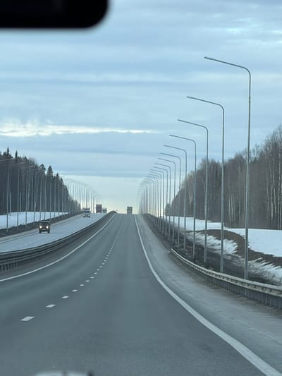 The scene depicts a multi-lane, divided asphalt highway in Khmelevka, Russia, viewed from inside a vehicle, indicated by a blurred dark object in the upper central frame. The highway ascends gently into the distance under an overcast sky characterized by layered grey-blue clouds. White solid and dashed lane markings are clearly visible on the grey pavement.

Parallel rows of grey metallic streetlights with curved arms and linear fixtures line both sides of the roadway, extending into the vanishing point. To the sides of the highway, patchy snow covers the ground, intermingled with exposed brown earth and sparse vegetation, consistent with late winter or early spring conditions. Dense forest, composed of both bare deciduous and evergreen trees, flanks the road. Metal guardrails, appearing as a concrete barrier on the left and a corrugated metal barrier on the right, delineate the edge of the roadway.

Several vehicles are in motion on the highway. In the opposing lanes on the left, a dark-colored passenger vehicle with illuminated headlights is visible in the leftmost lane, followed by two larger trucks further ahead on the incline. In the lanes traveling in the same direction, a light-colored truck precedes a larger, darker truck, both ascending the road in the distance. No human figures are discernible.